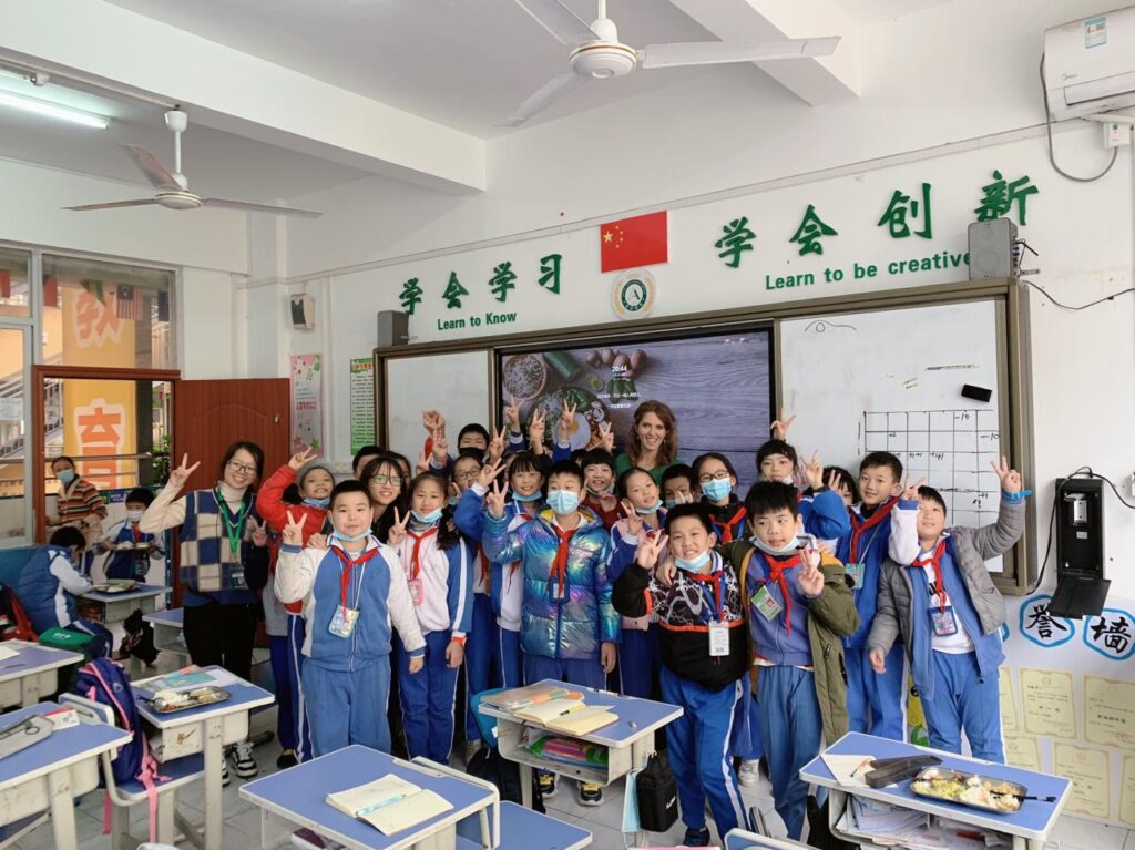 Teacher standing with a group of students in a classroom in China with Chinese and English signage visible on the walls.