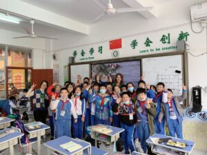 Teacher standing with a group of students in a classroom in China with Chinese and English signage visible on the walls.